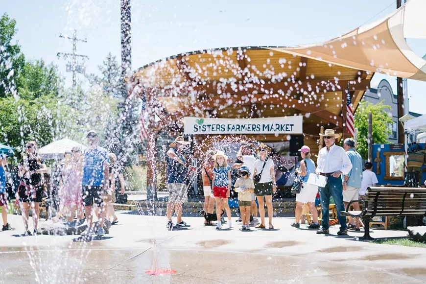 A sunny summer scene at Sisters Farmers Market shows families and visitors gathered around a splash pad in front of the market pavilion. Water sprays across the foreground as children play and adults watch, with the &ldquo;Sisters Farmers Market&rdquo; sign visible in the background under shade sails and blue skies.